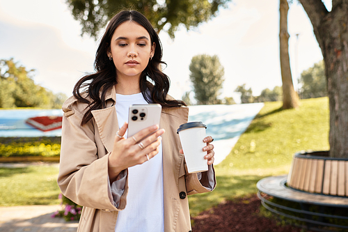 A young woman dressed warmly stands in a park, sipping coffee as she checks her phone.