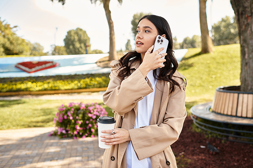 In a vibrant park, a young woman in cozy autumn attire chats on her phone, holding a warm drink.