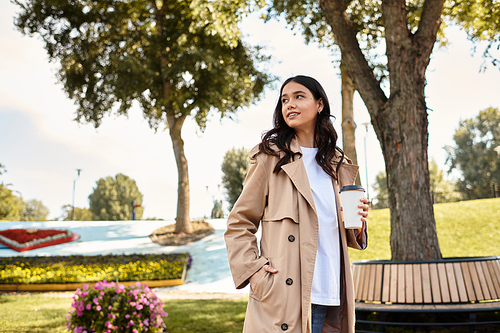 A young woman in stylish warm attire strolls through a vibrant park, sipping a warm drink.