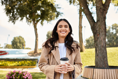 Dressed in warm autumn attire, a young woman smiles while holding a coffee cup in the park.