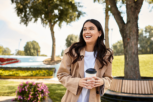 A cheerful young woman dressed in cozy autumn attire smiles while sipping her warm drink outdoors.