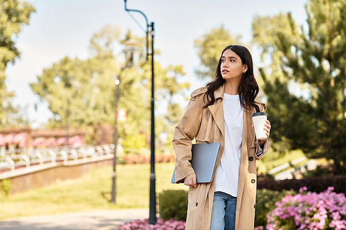 A beautiful young woman in autumn attire walks through a lush park, holding coffee and laptop.