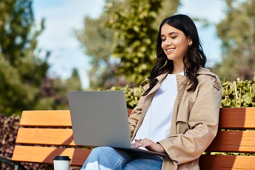 A young woman sits on a bench in a vibrant park autumn, focused on her laptop screen.