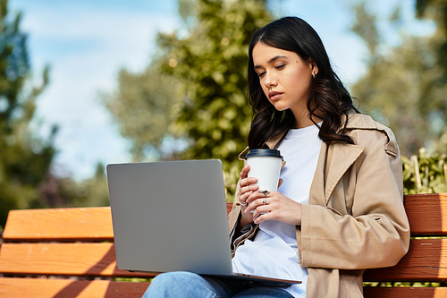 The beautiful young woman in cozy autumn attire is focused on her laptop while sipping a warm drink.