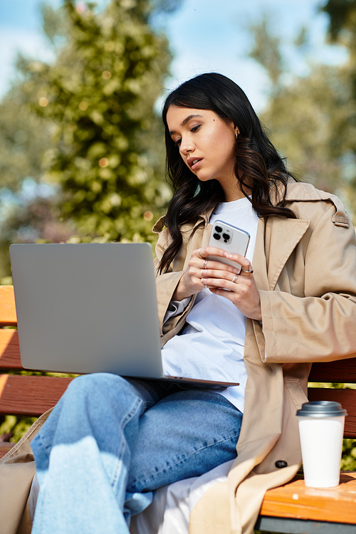 A young woman in autumn attire sits on a bench, focused on her laptop while sipping coffee.