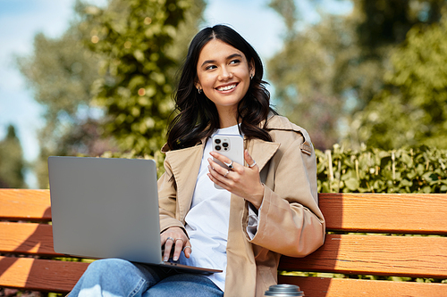 A cheerful young woman sits on a bench in the park, engaged with her laptop and smartphone.