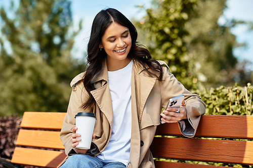 A young, beautiful woman in cozy fall attire smiles while checking her phone, surrounded by nature.