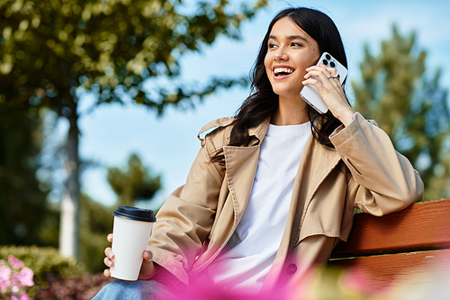 In the park, a young woman dressed warmly chats happily on her phone while enjoying coffee.