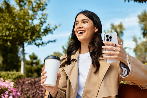 A young woman in autumn attire smiles with coffee and her phone outdoors.