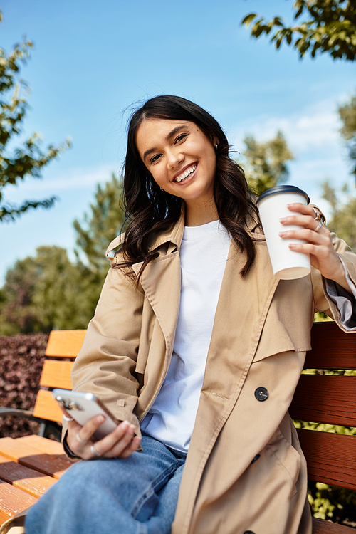 A cheerful woman in cozy attire relaxes on a park bench while sipping her coffee.