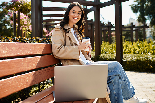 A young woman sits on a bench in the park, relaxing with coffee and focused on her laptop.