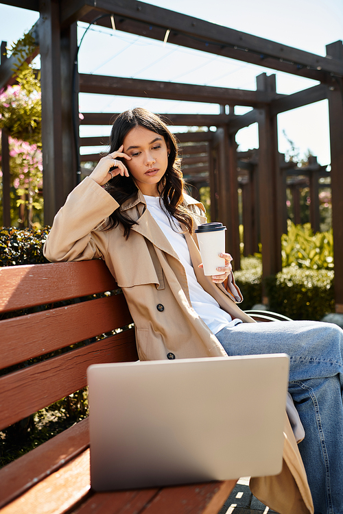 A stylish young woman sits on a park bench, sipping coffee while focused on her laptop.