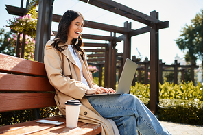 A young woman in warm attire sits on a bench, happily typing on her laptop in a peaceful park.