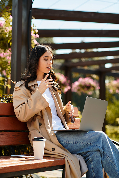 A young woman in warm attire sips coffee while using her laptop in a peaceful park.