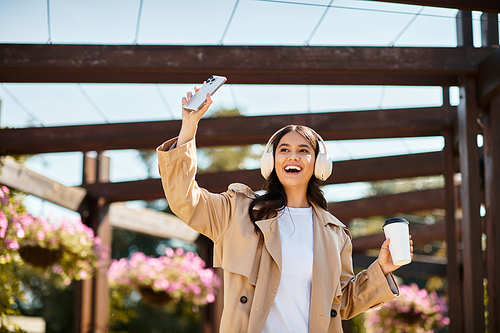 A cheerful woman wearing cozy autumn attire dances in the park, holding her phone and coffee.