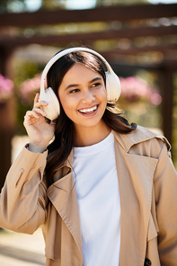 A young woman wears warm autumn clothing and headphones, smiling in a park.