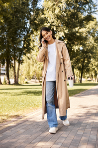 A young woman in warm clothes strolls through a scenic park, happily chatting on her phone.