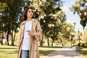 Stylishly dressed in autumn layers, a woman strolls through a sunlit park with coffee in hand.