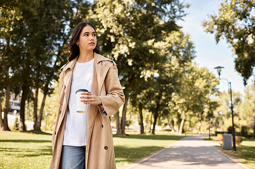 Stylishly dressed in autumn layers, a woman strolls through a sunlit park with coffee in hand.