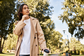 A young woman in autumn clothes pauses in a park to enjoy her coffee amid nature.