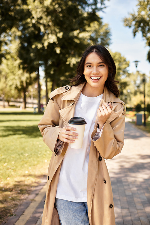 A young woman in cozy autumn attire radiates happiness while holding a warm drink in the park.