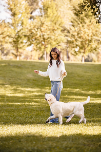 A young woman in autumn attire joyfully walks her dog through a lively park filled with trees.