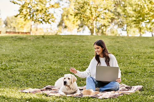 A young woman sits on a blanket in a park, engaged with her laptop while her dog relaxes beside her.