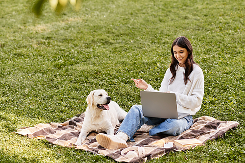 A cheerful young woman in warm autumn attire relaxes on a blanket with her dog, working outdoors.