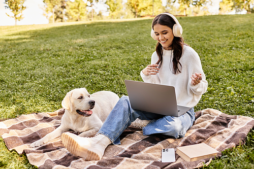 A young woman in warm attire sits on a blanket, joyfully using her laptop with a dog nearby.