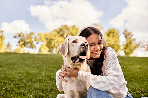 A young woman, wrapped in warm autumn clothing, embraces her dog while relaxing in the park.