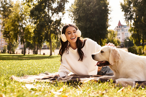 A cheerful young woman relaxes on grass, happily interacting with her dog in a vibrant park.