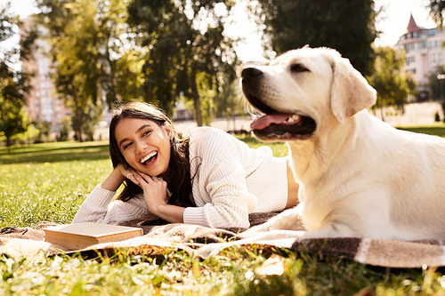 A beautiful woman relaxes on a blanket in the park, smiling while her dog lies beside her.