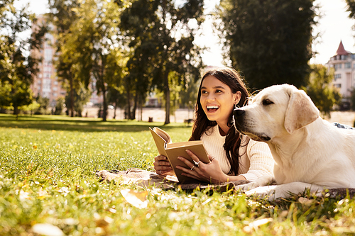 A young woman in cozy autumn attire sits on grass, happily reading with her dog beside her.