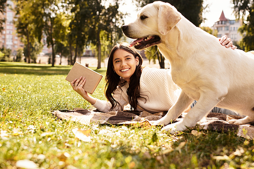 In a beautiful park, a young woman relaxes on a blanket, reading while her dog curiously watches.