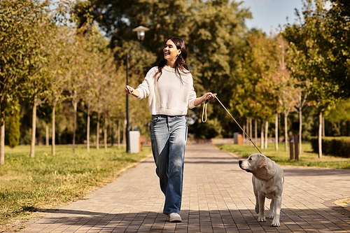 Wearing warm attire, a young woman joyfully strolls with her dog through a sunny park in autumn.