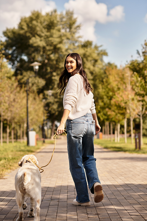 A joyful woman dressed in cozy autumn attire strolls through the park, holding her dogs leash.