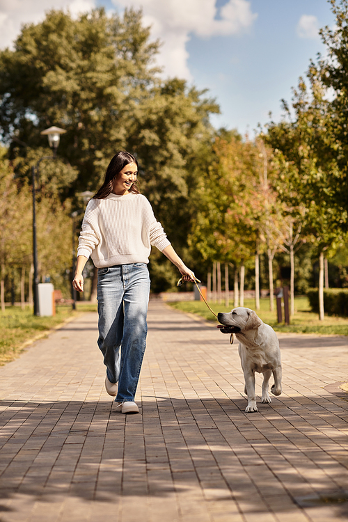 A young woman in warm attire strolls happily with her dog along a tree lined path in autumn.