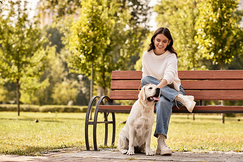 Delighted woman in cozy autumn wear interacts with her playful dog on a sunny park bench.