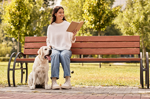 A young woman in warm attire smiles while reading in a sunlit park with her dog nearby.