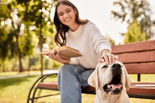 In a vibrant park, a young woman in cozy attire lovingly pets her smiling dog, immersed in nature.