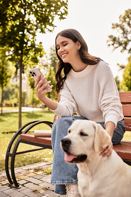 Sitting on a bench in the park, a young woman smiles while petting her dog and using her phone.