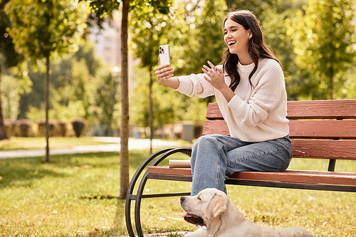 A young woman in cozy autumn attire smiles as she chats on her phone, seated in a park.