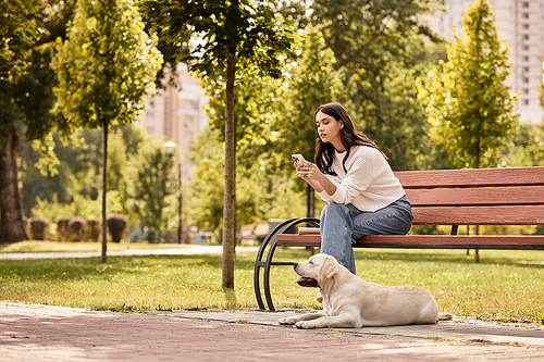 Sitting on a bench, a woman in cozy autumn attire looks at her phone while her dog relaxes nearby.