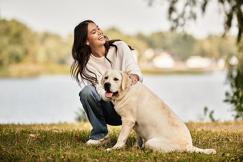 A woman in cozy autumn attire smiles warmly as she plays with her dog in a park setting.