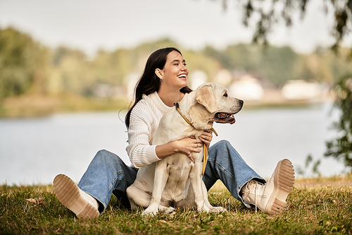 A young woman dressed in cozy autumn attire sits on the grass with her dog, smiling joyfully.