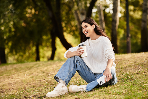 A young woman in warm clothes happily sips coffee in a park filled with autumn leaves.