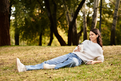 A young woman lounges in the park, sipping a warm drink while surrounded by autumn foliage.