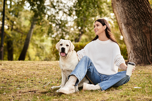 A young woman in cozy autumn attire relaxes on the grass with her cheerful dog, sipping coffee.