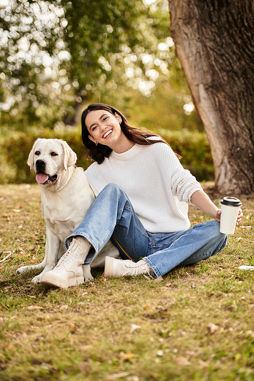 A young woman in cozy autumn attire sits happily on the grass beside her dog, sipping coffee.