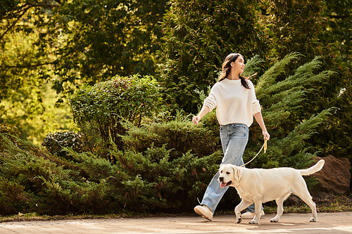 A young woman in cozy autumn attire walks joyfully with her happy dog amidst falling leaves.